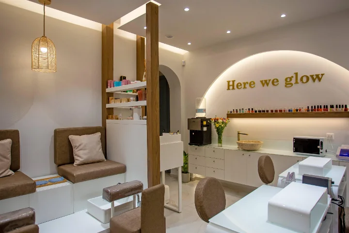 Un salone unghie caldo e dalla luce soffusa, con una lampada a sospensione in rattan intrecciato, panche pedicure marroni imbottite, tavoli manicure bianchi, una mensola di smalti colorati e un'insegna dorata «Here we glow» su un arco retroilluminato.