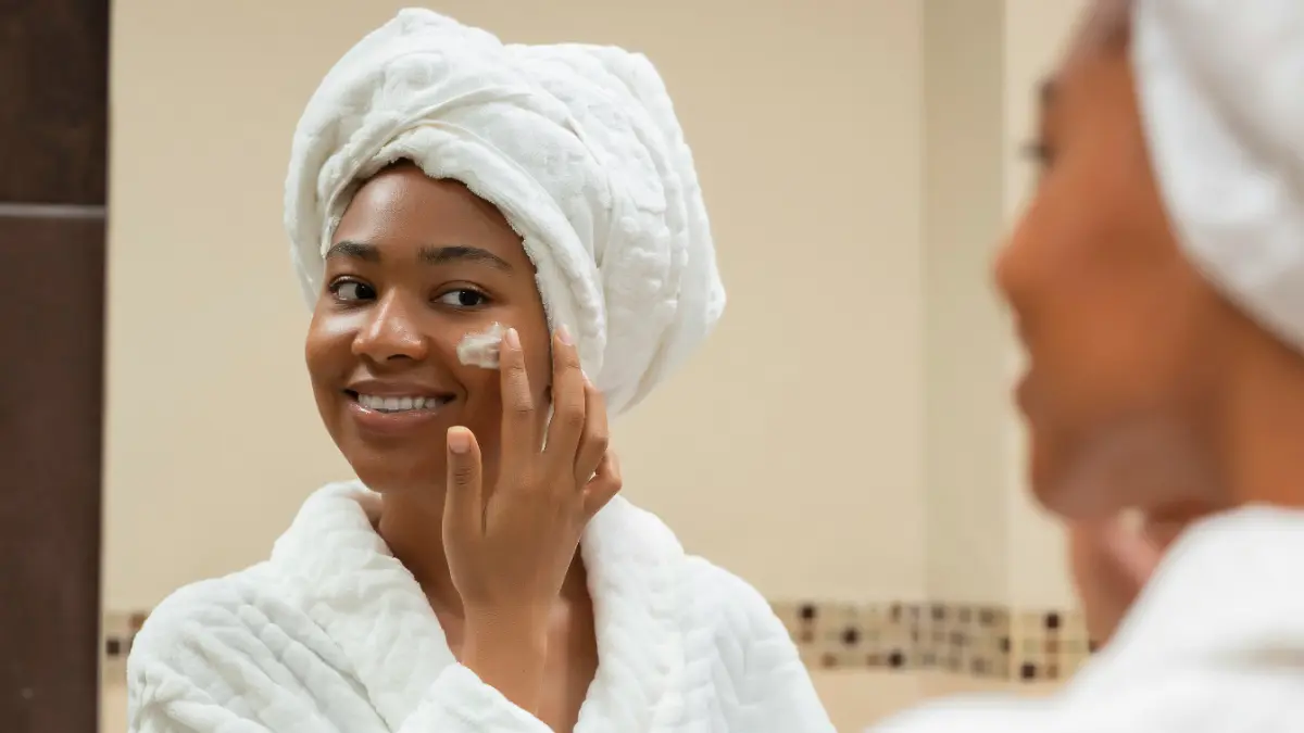 Una donna in accappatoio bianco con un asciugamano bianco avvolto intorno alla testa sorride allo specchio del bagno mentre applica l'SPF sul viso
