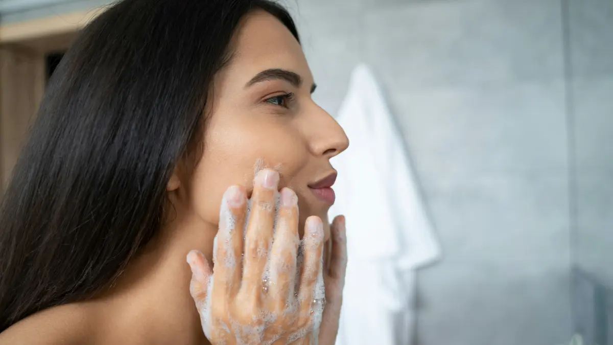 Vista di lato di una donna che massaggia un detergente sul viso, chinata sul lavandino del bagno