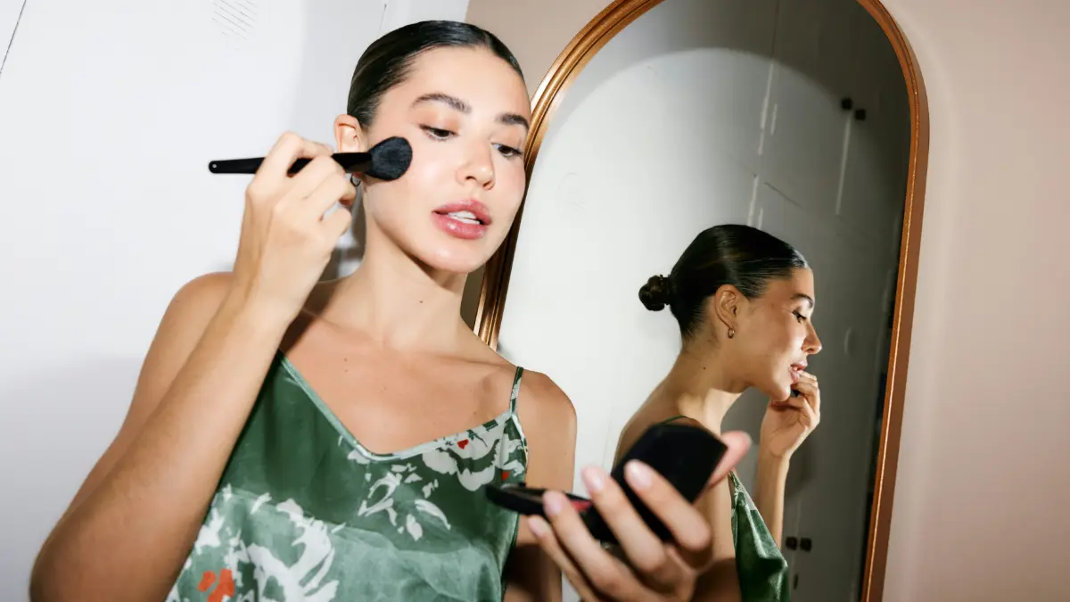 Una donna elegante e pronta per uscire si guarda in uno specchietto mentre applica il make-up con un pennello