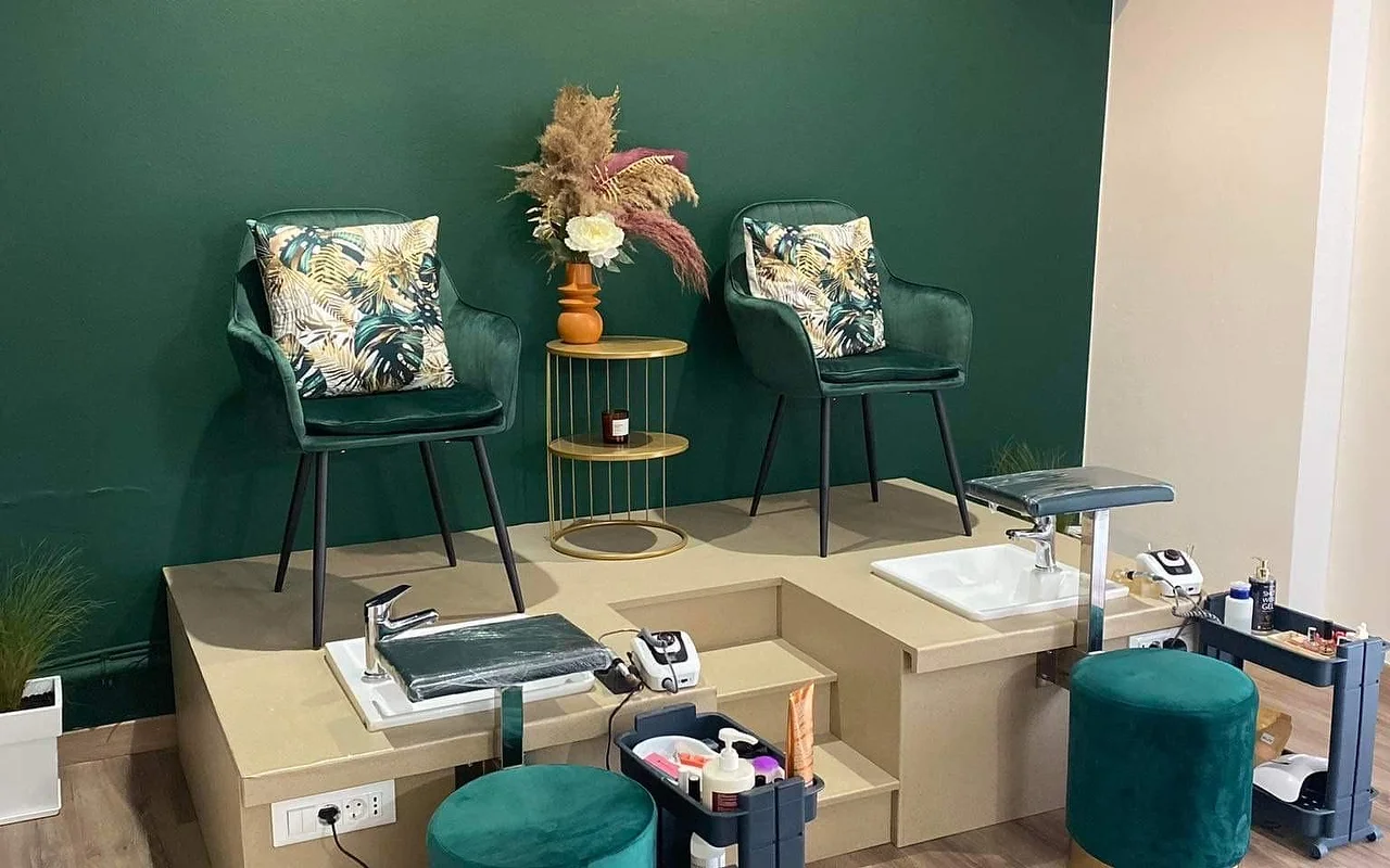 Un angolo pedicure con una parete verde intenso, due poltrone a botte in velluto verde con cuscini a stampa tropicale rialzate su una piattaforma beige sopra vasche pedicure, un tavolino dorato con pampas in un vaso arancione e sgabelli abbinati in velluto verde.