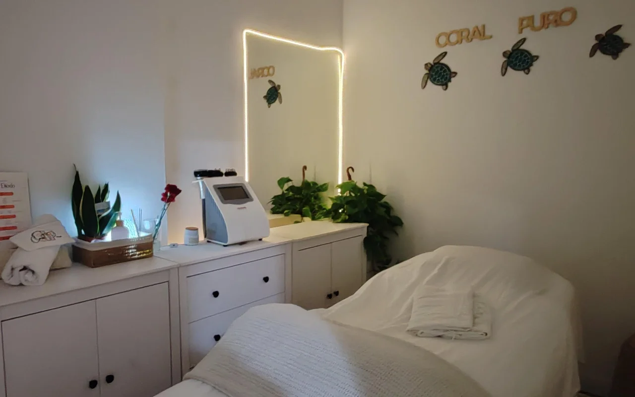 Una piccola e tranquilla sala trattamenti con armadietti bianchi e un lettino apparecchiato con asciugamani bianchi. Uno specchio illuminato e un macchinario per trattamenti stanno sul bancone tra piante verdi, sotto la scritta dorata «Coral Puro» e un'arte murale decorativa a forma di tartaruga.