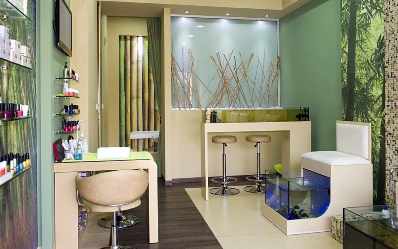 Un salone compatto con decorazioni murali in bambù e verde foresta, mensole a parete allineate di smalti per unghie, un tavolo manicure e sedia color crema, due sgabelli da bar e una vasca di vetro illuminata di blu per la pedicure fish spa.