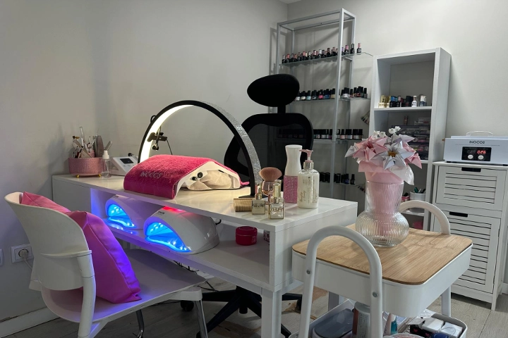 Postazione singola manicure con scrivania bianca, una ring light e due lampade LED con luce blu, una sedia nera e mensole ricoperte di smalti colorati, ravvivate da un vaso rosa pieno di fiori.