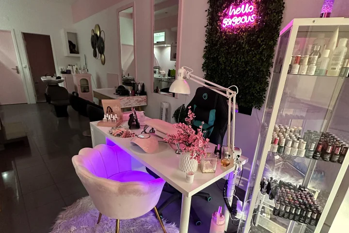 Postazione beauty illuminata di rosa, con un'insegna al neon 'hello gorgeous' su una parete vegetale, una poltrona crema a forma di conchiglia, una scrivania bianca con pennelli e fiori di ciliegio, e una vetrina piena di smalti colorati.