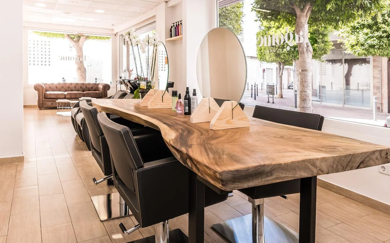 Un salone di capelli luminoso costruito attorno a un lungo bancone in legno con bordo naturale, allineato con poltrone da parrucchiere nere. Uno specchio rotondo e supporti in legno per i prodotti stanno sul bancone, un divano Chesterfield in pelle marrone attende sul fondo, e alte finestre danno su una strada alberata.