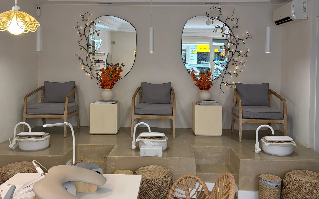 Un salone per unghie moderno e luminoso con due sedie da pedicure su una piattaforma di cemento rialzata, ciascuna con una bacinella bianca per i piedi e una poltrona grigia imbottita. Due specchi dalle forme organiche, decorati con rami luminosi e fiori arancioni, sono appesi sopra, con una postazione manicure in primo piano.