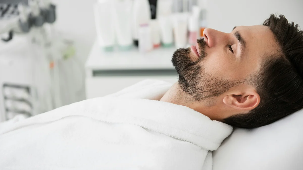 Un uomo è sdraiato sul lettino con un accappatoio bianco, la testa reclinata all’indietro e gli occhi chiusi