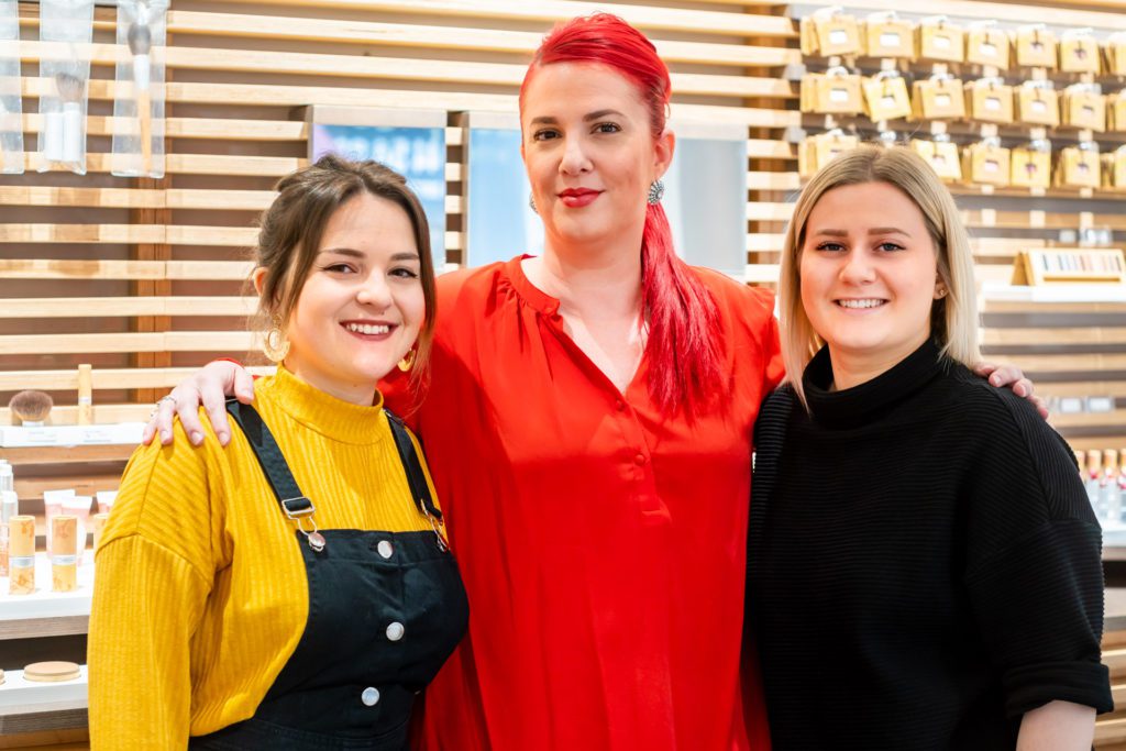 Drei lächelnde Salon-Mitarbeiterinnen posieren Arm in Arm vor einer Holzlattenwand, an der Kosmetikprodukte präsentiert werden. Die Frau in der Mitte hat leuchtend rotes Haar und ein rotes Oberteil.