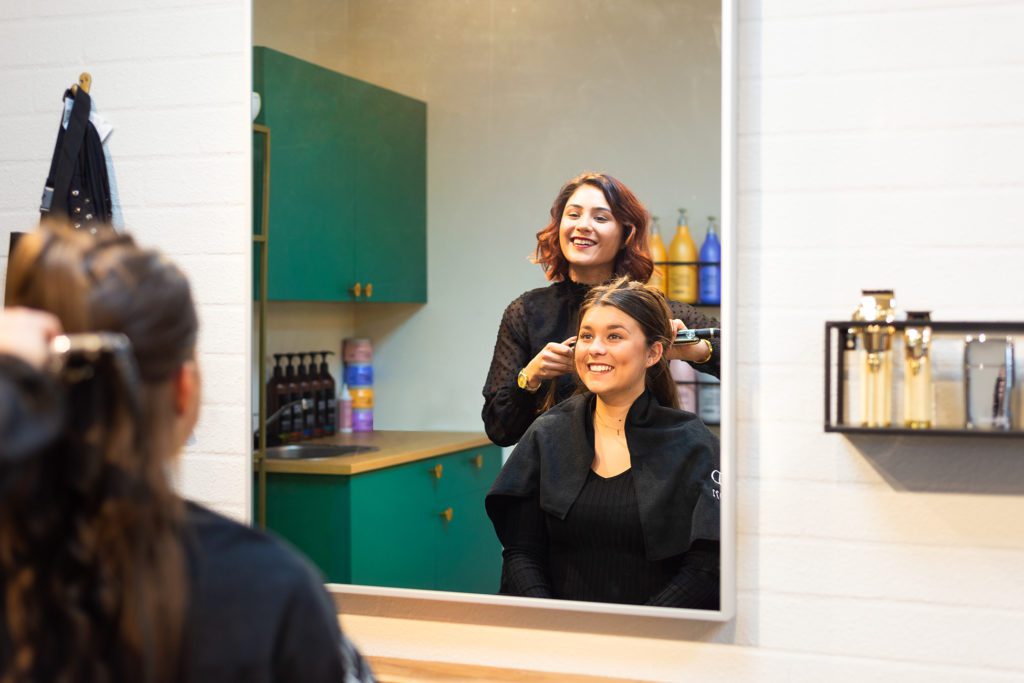 Eine lächelnde Friseurin mit kastanienbraunem Haar stylt das Haar einer lächelnden Kundin mit einem Glätteisen, gesehen durch das Spiegelbild eines grossen rechteckigen Spiegels in einem modernen Salon mit smaragdgrünen Schränken.