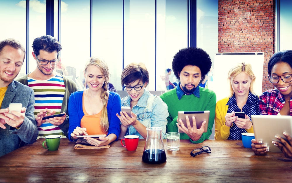 Vielfältige Gruppe von Menschen an einem Tisch, die Smartphones und Tablets nutzen – sinnbildlich für die Online-Weiterbildung von Coiffeuren und Barbieren