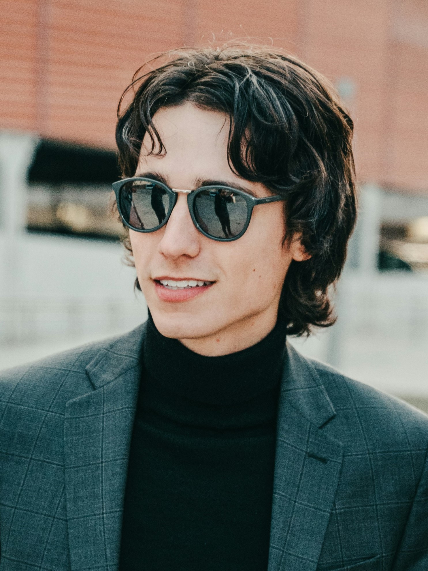 Ein junger Mann mit Retro-Vokuhila, der ein graues Sakko und Sonnenbrille trgt und draussen lchelnd steht.