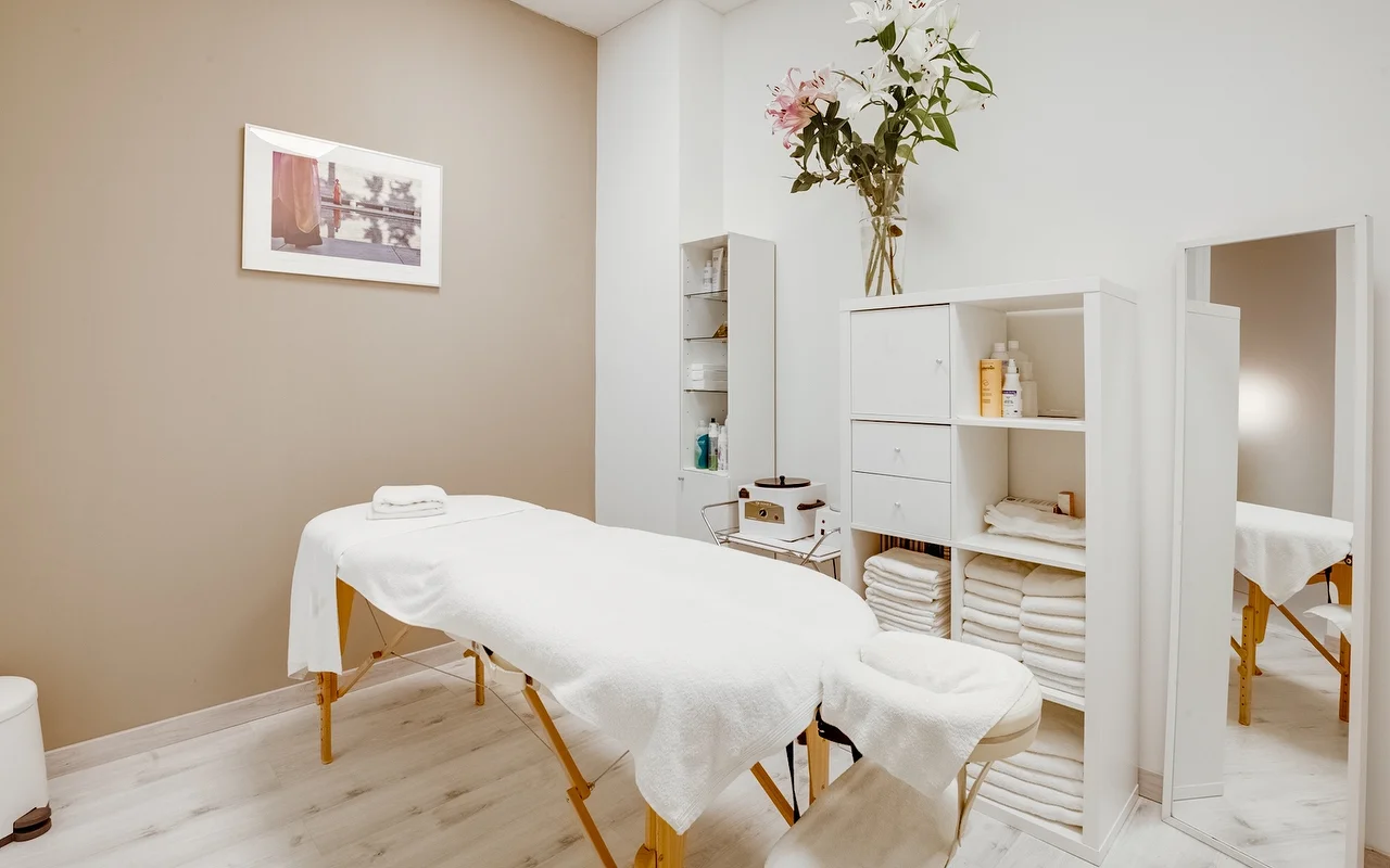 Ein ruhiger Behandlungsraum mit einer weissen Massageliege und frischen Handtüchern. Der Raum hat neutrale Beige- und Weisstöne, einen weissen Schrank mit gefalteten Handtüchern und eine hohe Glasvase mit weissen und rosafarbenen Lilien.