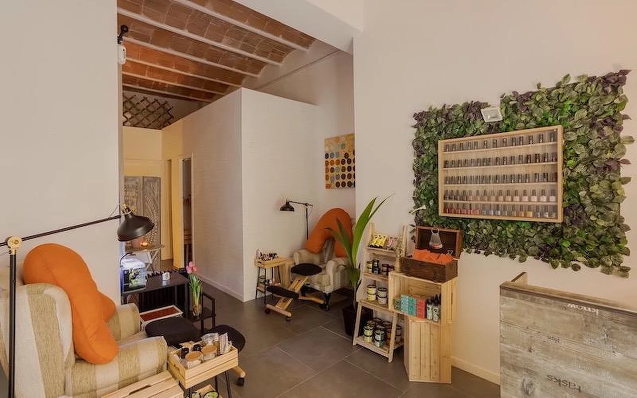 Ein gemütliches Nagelstudio im Bohème-Stil mit weichen Sesseln und orangefarbenen Kissen. An der Wand eine grosse vertikale Pflanzeninstallation rund um ein Holzregal voller Nagellackflaschen.