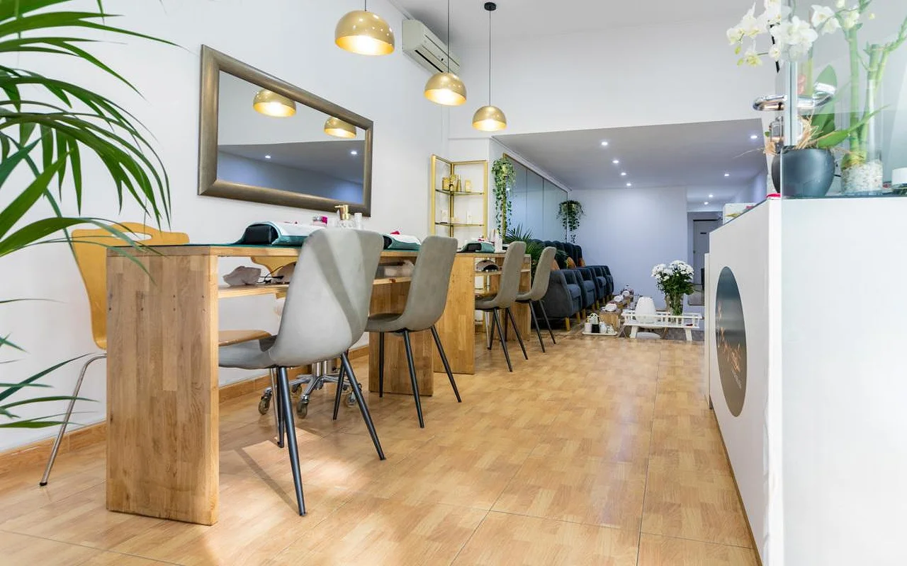 Ein helles Nagelstudio mit langen Holz-Maniküretischen und grauen modernen Stühlen. Goldene Kuppel-Pendelleuchten hngen über den Pltzen, und ein grosser Spiegel mit dunklem Rahmen hngt an der weissen Wand.