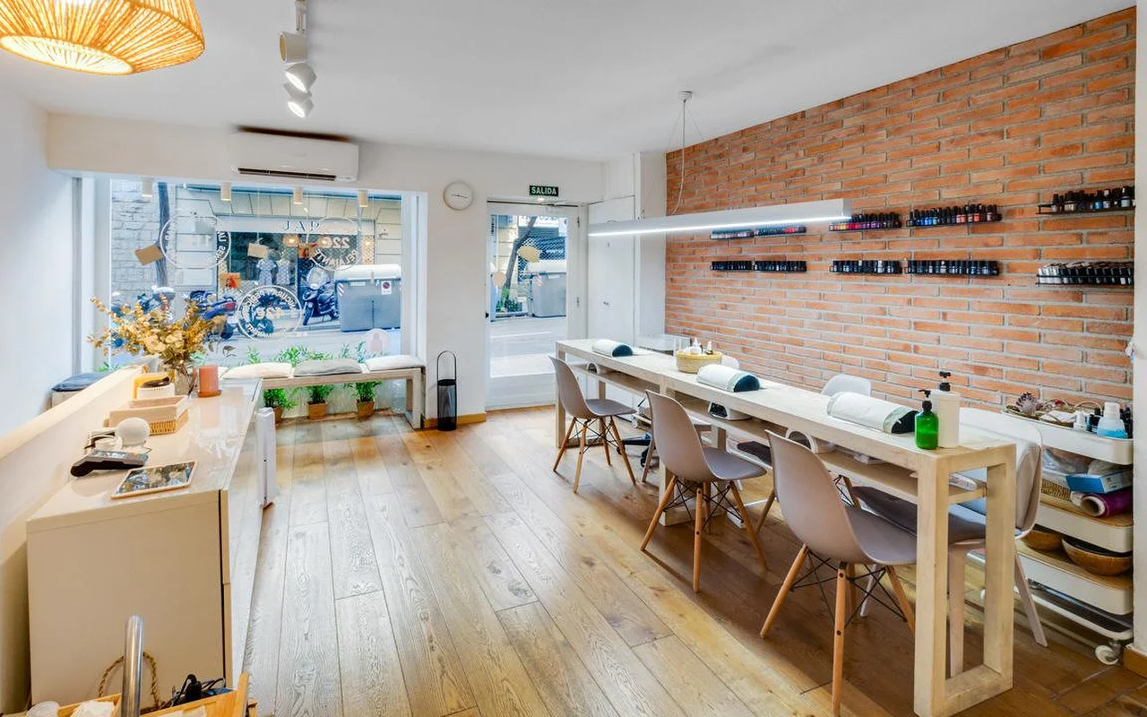 Ein moderner Salon mit rustikalem Akzent: eine lange weisse Maniküre-Theke und eine rote Backsteinwand. Grosse Fenster blicken auf eine Stadtstrasse, und der Holzboden wird von natürlichem Licht durchflutet.