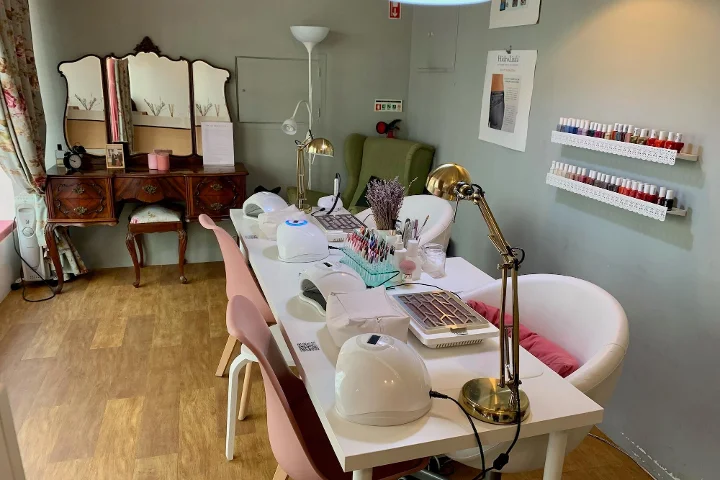 Innenansicht eines gemütlichen Schönheitssalons mit Schwerpunkt Maniküre. Im Vordergrund ein langer weisser Tisch mit Nagellampen, Schreibtischleuchten und verschiedenen Nageldesign-Werkzeugen, flankiert von rosa und weissen Stühlen. Im Hintergrund ein Vintage-Schminktisch mit dreiteiligem Spiegel, ein grüner Sessel und weisse Wandregale mit Reihen bunter Nagellackflaschen.