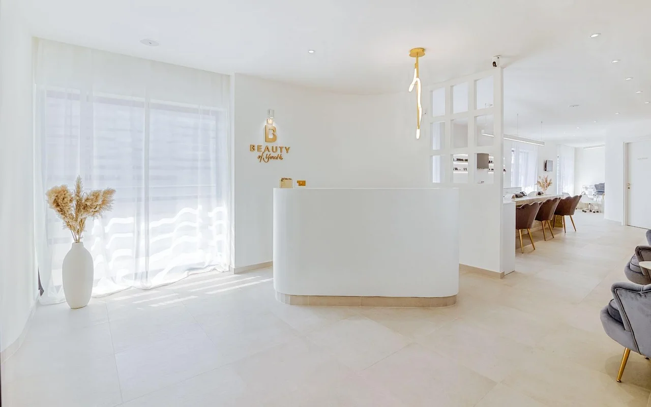 Ein heller, ganz in Weiss gehaltener Empfangsbereich mit geschwungenem Tresen, goldenem Beauty-of-Youth-Logo an der Wand, einer skulpturalen Hängelampe, durchscheinenden Vorhängen und braunen Samt-Behandlungsstühlen im Hintergrund.