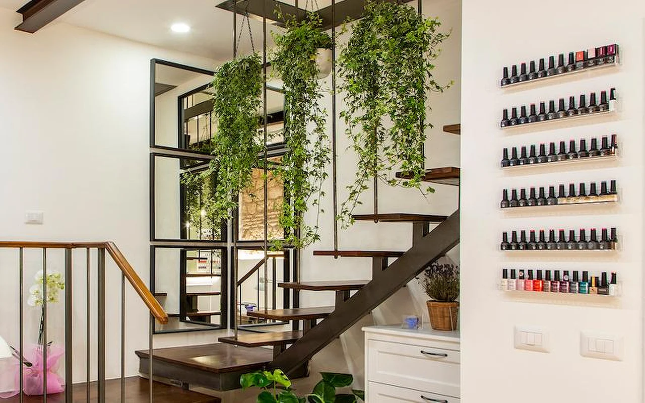 Ein stilvolles, modernes Interieur mit einer freitragenden Treppe aus Holz und Metall. Üppige Hängepflanzen ranken von der Decke zwischen den Stufen, und Reihen bunter Nagellackflaschen stehen auf schlanken, transparenten Regalen an der weissen Wand.