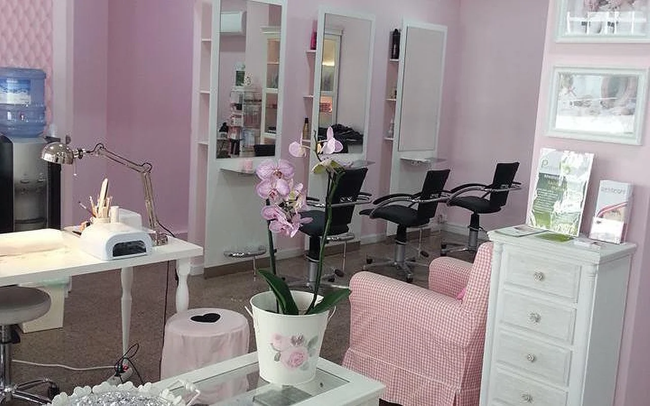 Das Innere eines hellen, pinkfarbenen Haar- und Nagelsalons. Links eine weisse Manikürestation mit Schreibtischlampe und UV-Gerät. In der Mitte eine Orchidee in einem weissen Topf auf einem Couchtisch neben einem rosa-weiss karierten Sessel. Im Hintergrund mehrere Frisierstationen mit hohen Spiegeln und schwarzen Salonstühlen vor hellrosa Wänden.