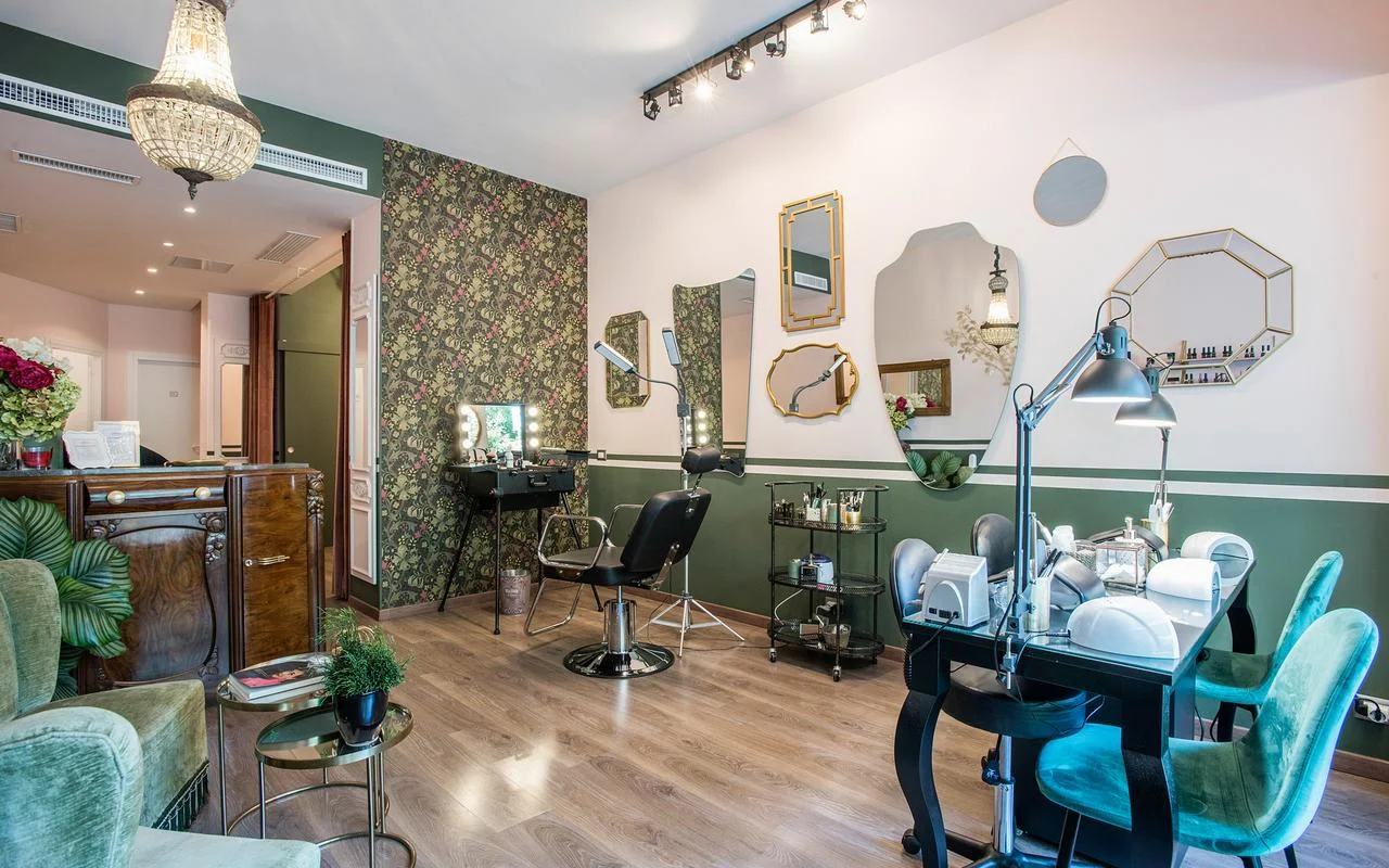 Ein Salon im Vintage-Chic-Stil mit weissen und waldgrünen Wänden. Blumentapete, eine Sammlung unregelmässig geformter, goldgerahmter Spiegel, eine professionelle Make-up-Station und üppige grüne Samtsitze.