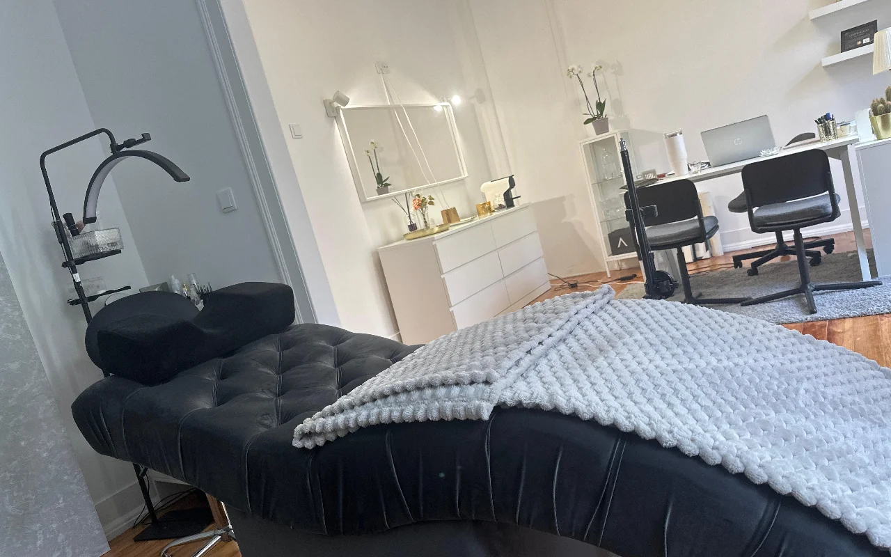 Ein moderner Raum für Wimpernbehandlungen aus einer niedrigen Perspektive. Im Vordergrund eine gepolsterte schwarze Beauty-Liege mit einer strukturierten grauen Decke und einem ergonomischen Nackenkissen unter einem gebogenen Ringlicht. Im Hintergrund ein minimalistischer weisser Bürobereich mit Schreibtisch, zwei schwarzen Stühlen, Laptop, weisser Kommode und grossem Spiegel.