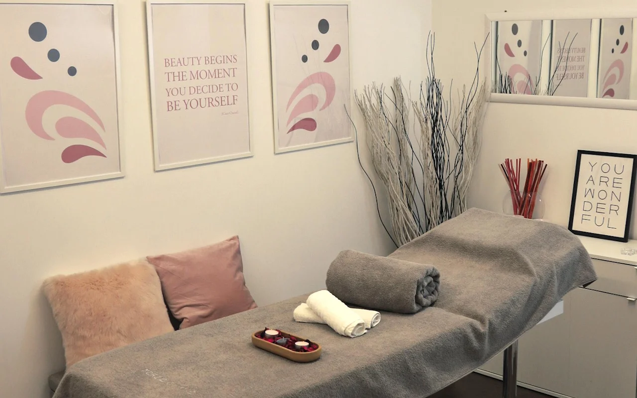 Ein heller Behandlungsraum mit einer Massageliege, die mit einem grauen Handtuch bedeckt ist. Die Wand ist mit abstrakter Kunst in Pastelltönen und motivierenden Sprüchen dekoriert, während dekorative weisse Zweige und Kerzen eine ruhige Atmosphäre schaffen.