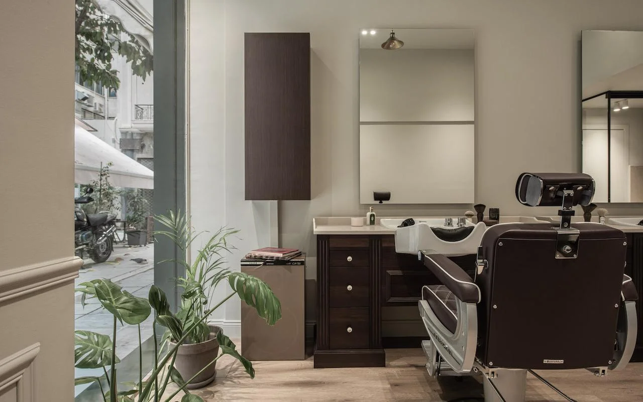 Ein stilvoller Barbershop mit dunkelbraunem Leder-Friseurstuhl, Schrank aus Walnussholz, einem grossen Fenster mit Blick auf eine Kolonaki-Strasse und einer Monstera-Pflanze, die dem minimalistischen Raum etwas Grün verleiht.