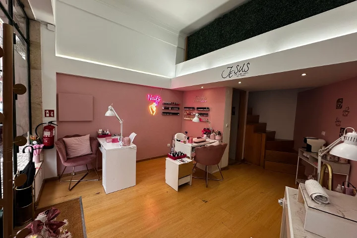 Ein schickes Nagelstudio mit hellrosa Wänden und warmem Holzboden. Der Raum enthält mehrere weisse Maniküretische mit altrosa Samtstühlen. Ein leuchtend pinkfarbenes Neonschild in Form einer Hand mit Nagellackflasche und dem Wort „Nails“ leuchtet an der Wand, und Reihen von Nagellacken stehen auf kleinen Regalen. Ein Zwischengeschoss mit grüner Kunsthecken-Wand ist oben sichtbar.