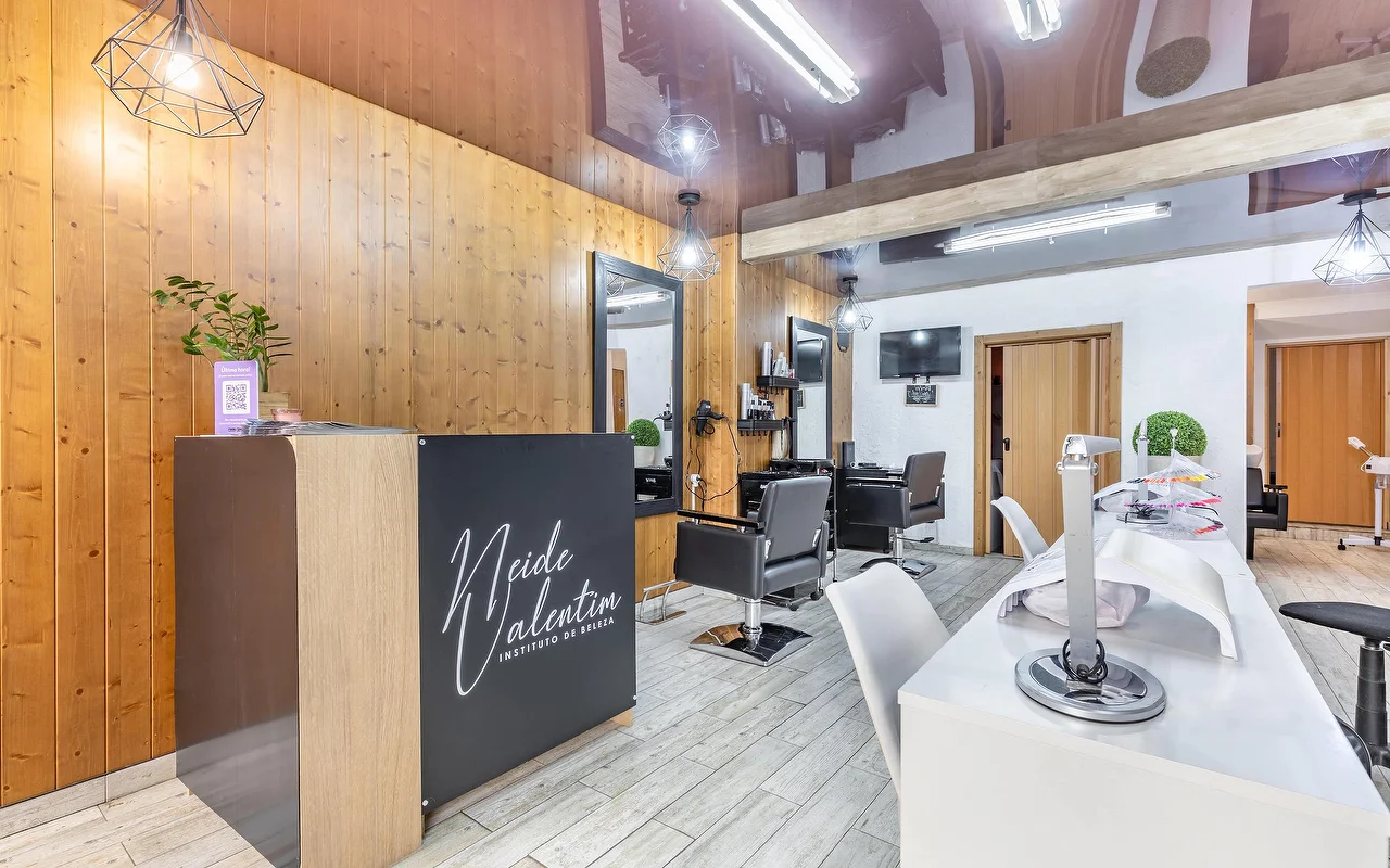 Ein moderner Schönheitssalon mit einem Empfangstresen links, beschriftet mit „Neide Valentim Instituto de Beleza“. Die Wände sind mit warmem, vertikalem Holz verkleidet, dazu grosse Spiegel. Schwarze Frisierstühle stehen vor den Spiegeln, während eine weisse Manikürestation mit weissen Stühlen an der rechten Seite des grau gefliesten Bodens verläuft.