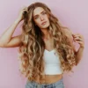 Studioaufnahme einer Frau mit sehr langem, welligem, sonnengeküsstem Balayage-Haar, die eine hellblaue Jeans und ein weisses Strick-Crop-Top trägt
