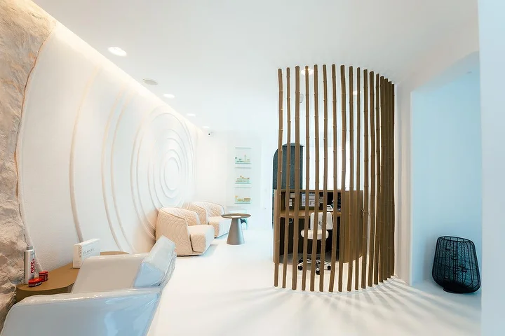Ein ruhiger, höhlenartiger Spa-Innenraum mit geschwungenen weissen Wänden, eine davon in konzentrischen Wellen ausgearbeitet, sanftes indirektes Licht, cremefarbene Steppsessel und ein geschwungener Holzlamellen-Paravent, der eine ruhige Behandlungsnische abtrennt.