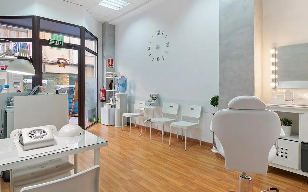 Ein heller, weisser Salon mit hellem Holzboden, einer Reihe weisser Wartestühle, einer minimalistischen Wanduhr, einem beleuchteten Schminkspiegel, einem weissen Behandlungsliegestuhl und einer Maniküre-Station am Fenster.