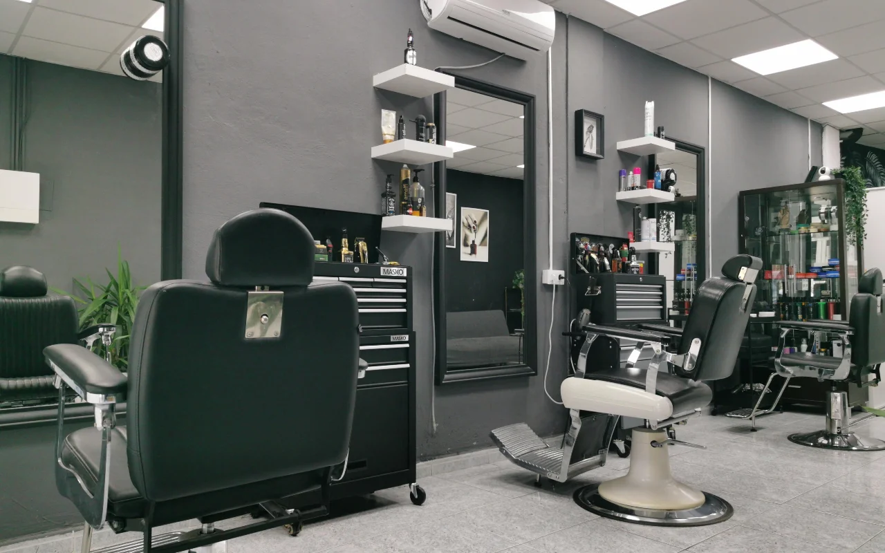 Ein traditioneller Barbershop mit anthrazitgrauen Wänden, klassischen schwarzen Lederbarberstühlen mit Chromfuss und Fussstützen, weissen schwebenden Regalen voller Pflegeprodukte und grossen Spiegeln auf gefliestem Boden.