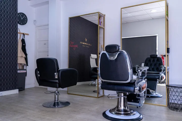 Ein reduziert eingerichteter Salon mit einem schwarzen Stylingstuhl und einem Barberstuhl aus Chrom und Leder auf hellem Holzboden, einem grossen goldgerahmten Spiegel, einer Wanduhr und Umhängen, die neben einer dunklen Akzentwand hängen.