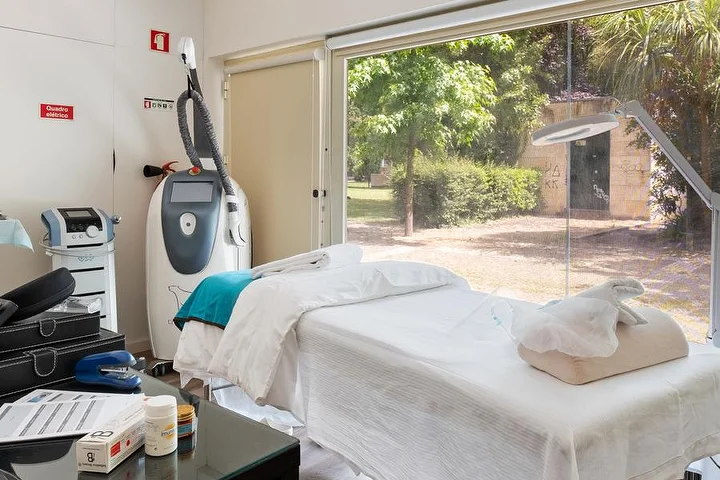 Ein Behandlungsraum mit einer weiss bezogenen Liege neben einem bodentiefen Fenster zu einem grünen Garten, einem Körperbehandlungsgerät auf einem Ständer, einer Lupenlampe und einem Glastisch mit Produkten.