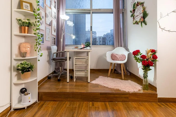 Ein kleines Kosmetikstudio auf einem erhöhten Holzpodest, mit einem weissen Schreibtisch und einer Klemmlampe, einem Bürostuhl und einem Sessel, Eckregalen mit Pflanzen, einer Vase mit roten Rosen und einem Fenster zur Stadt.