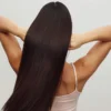Rückansicht einer Frau in einem weissen Trägertop, die mit erhobenen Armen durch ihr langes, glänzendes brünettes Haar fährt