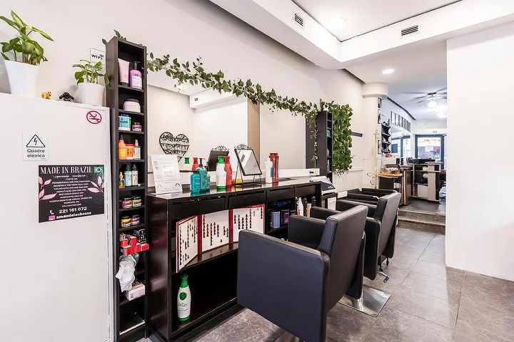 Ein schmaler Friseursalon mit schwarzen Stylingstühlen entlang einer verspiegelten Wand, einem dunklen Regal voller Haarpflegeprodukte, einer herabhängenden Grüngirlande über dem Spiegel und einem gerahmten Made-In-Brazil-Schild auf einer weissen Säule.
