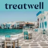 Die Uferpromenade von Klein-Venedig in Mykonos-Stadt: weiss getünchte Häuser mit bunten Holzbalkonen direkt am Wasser, dahinter die türkisfarbene Ägäis und im Vordergrund weisse und petrolfarbene Café-Tische auf einer steingepflasterten Terrasse.