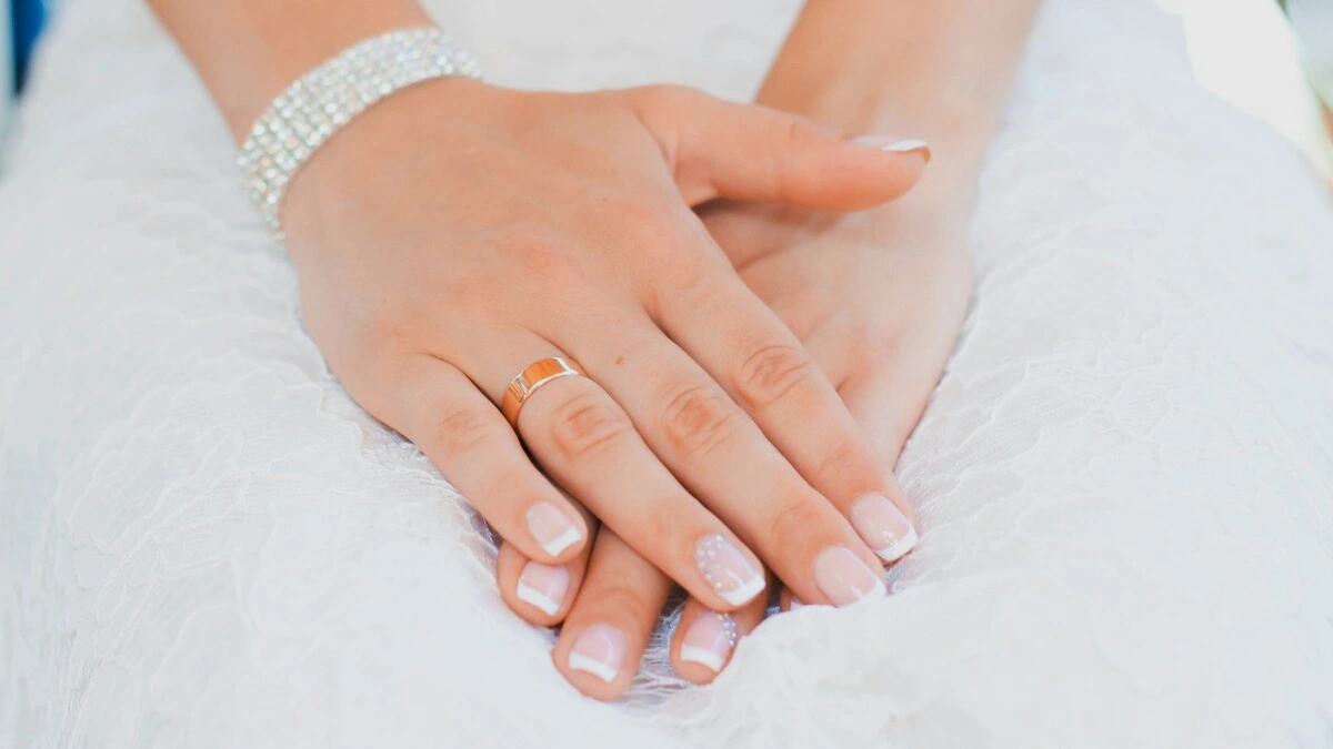Die Hände einer Frau ruhen in ihrem Schoss; sie trägt ein Spitzen-Hochzeitskleid und eine gepflegte French-Tips-Maniküre in klassischer Kombination aus Nude und Weiss auf kurzen, eckig-ovalen Nägeln, mit zarter Verzierung an den Ringfingern