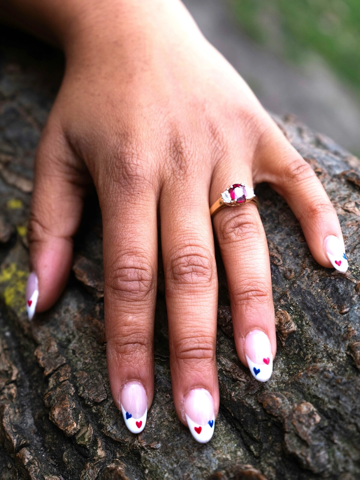 Die Hand einer Frau ruht auf einem Ast und zeigt eine French-Tips-Maniküre auf ovalen Nägeln, verziert mit kleinen roten und blauen Herzen