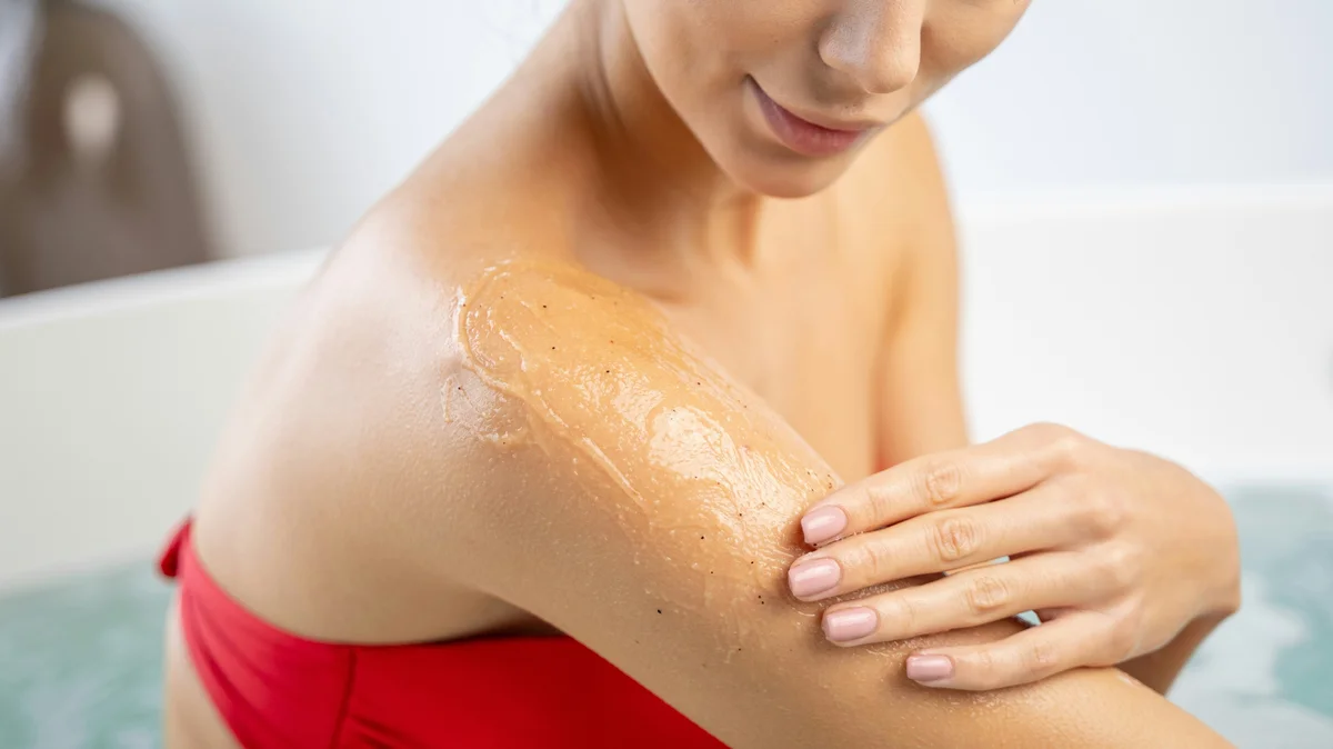 Nahaufnahme einer Frau in einem roten Bikini, die in einem sauberen, weissen Bad ein Peelinggel auf ihren Oberarm aufträgt