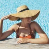 Eine Frau in einem sonnigen Aussenpool stützt sich mit den Armen am Beckenrand ab, sie trägt einen orangebraunen Bikini und einen breitkrempigen Strohhut