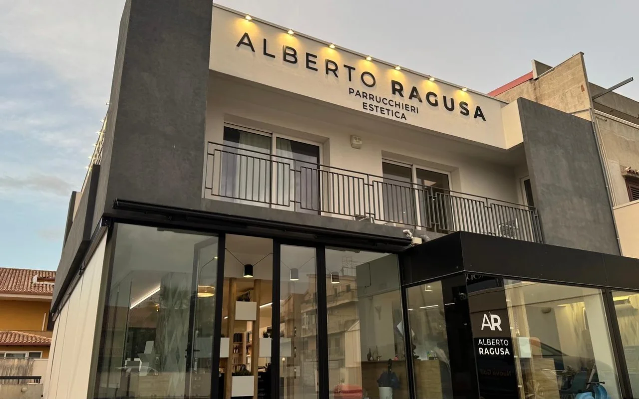 Das Äussere von Alberto Ragusa in der Dämmerung, ein zweistöckiger Salon mit einem grossen beleuchteten Schild „Alberto Ragusa Parrucchieri Estetica“ über raumhohen Glasfenstern, mit warmem Licht, das von innen strahlt.