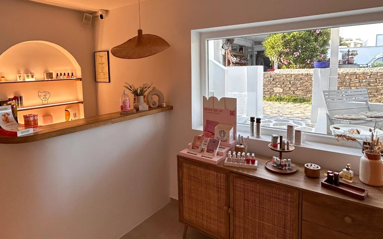 Das Innere von BeautiQ Esthetics and More auf Paros, mit einer sanft beleuchteten gewölbten Nische voller Nagellacke, einer geflochtenen Rattan-Hängeleuchte und einem grossen Fenster mit Blick auf eine Steinmauer und einen sonnigen Garten.