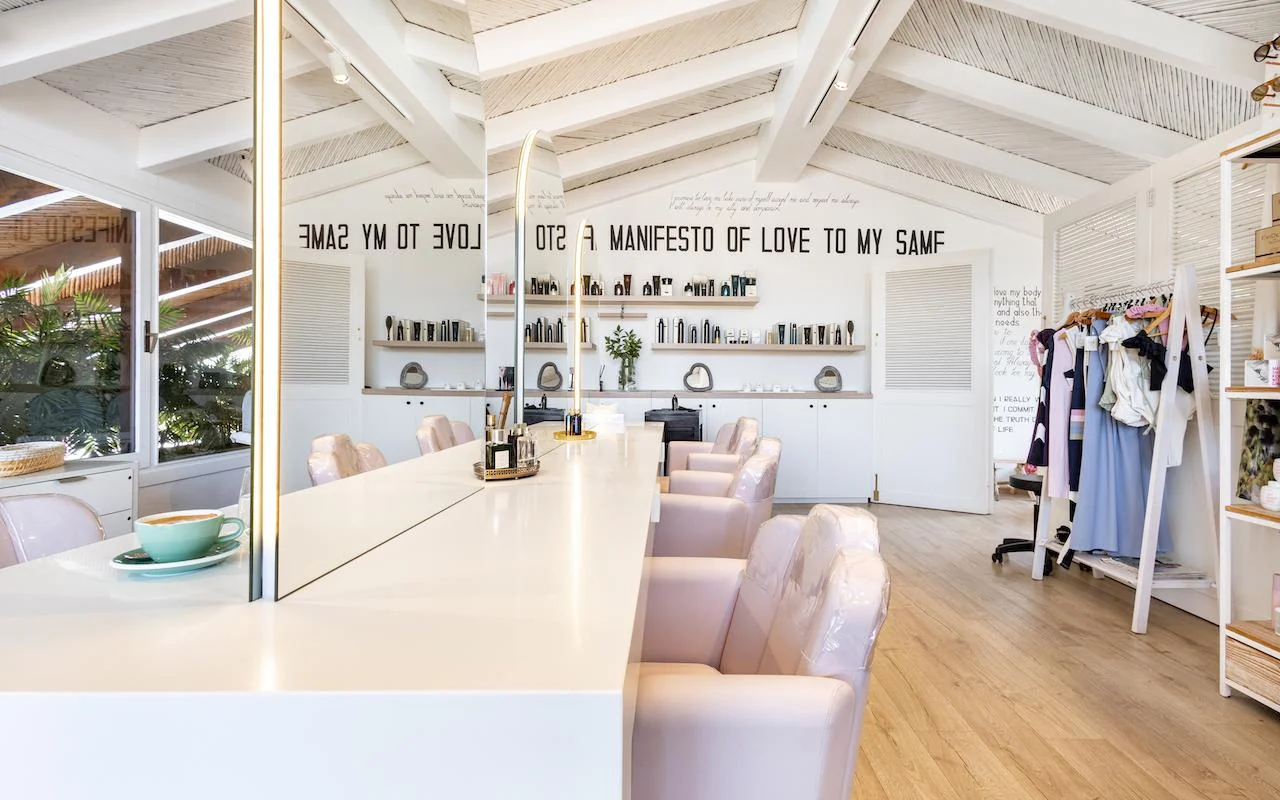 Das helle Interieur von Milk & Roses in Puente Romano, Marbella, mit einem weissen Styling-Tresen, blassrosa Stühlen, einer Balkendecke und Produktregalen entlang der Wand.