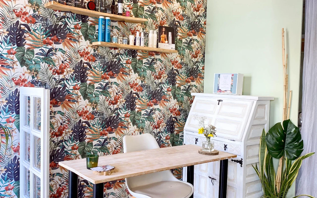 Ein Styling-Platz bei Shangara Lora Organics in Marbella, mit auffälliger tropischer Tapete, einem Holztisch, einem antiken weissen Sekretär und Bio-Haarpflege auf schwebenden Regalen.