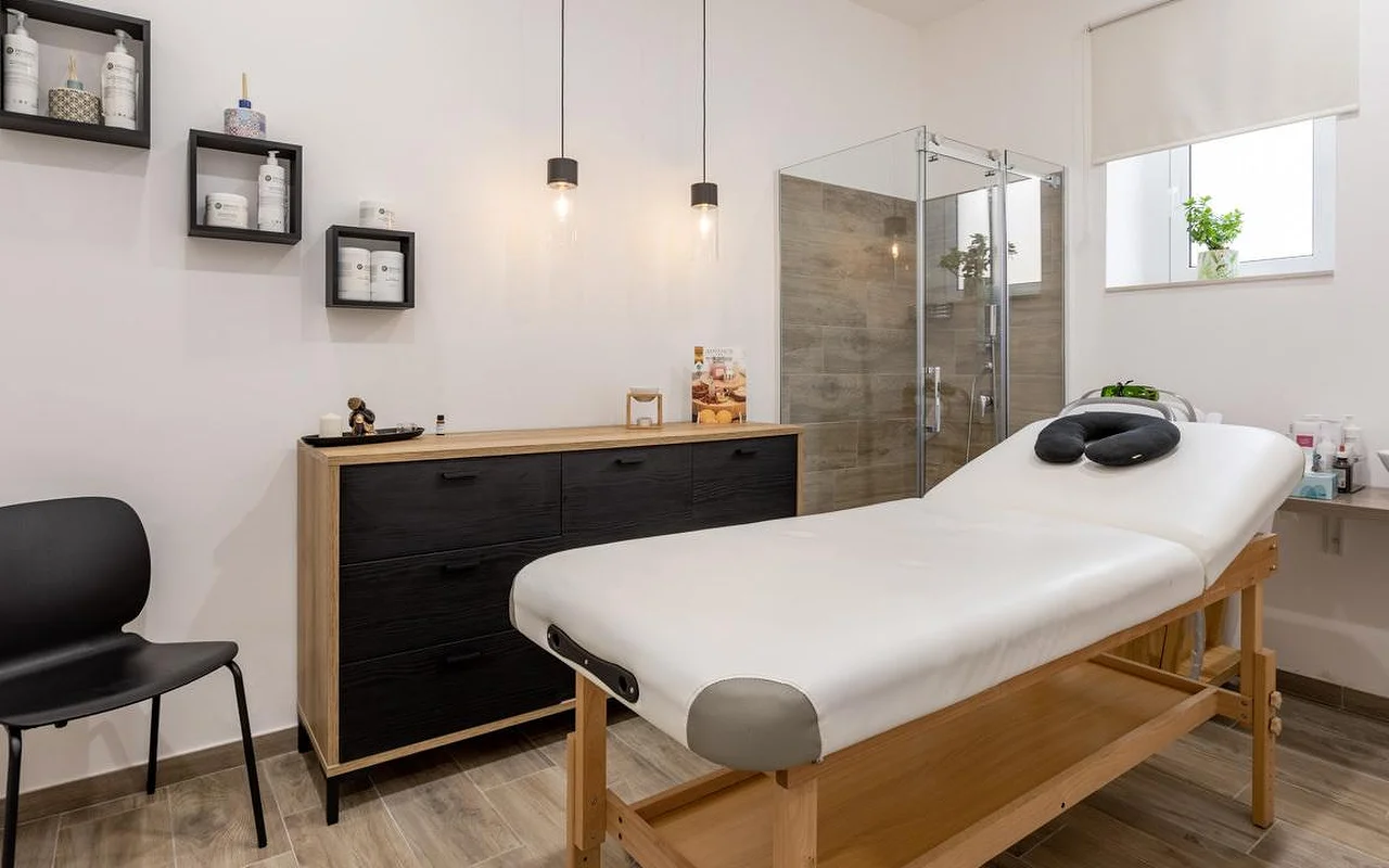 Ein Massageraum bei Thea Estetica, mit einem hölzernen Massagetisch mit schwarzer Gesichtsauflage, einer Kommode in Schwarz und Holz, zwei Pendelleuchten, Würfelregalen mit Produkten und einer Glasduschkabine mit Fliesen in Holzoptik.