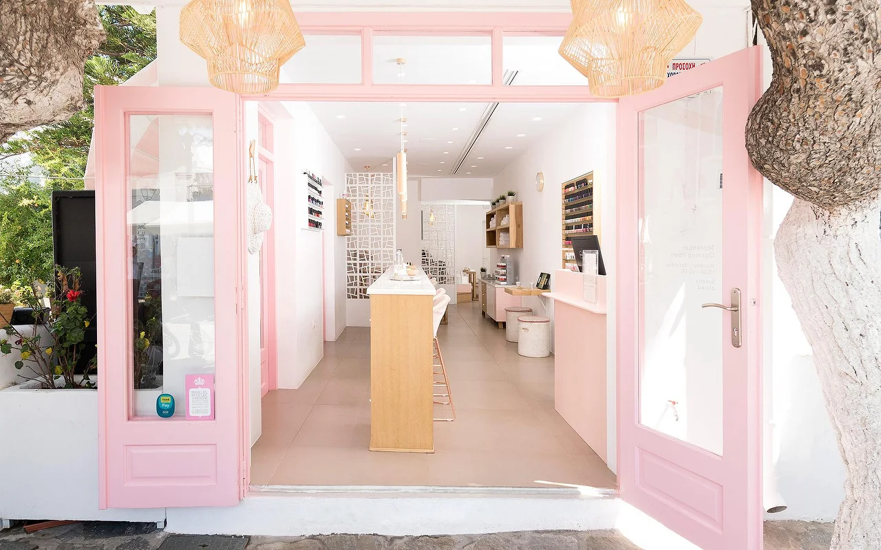 Der rosa gerahmte Eingang von Vivian Nail Boutique in Naoussa auf Paros, mit offenen Türen, geflochtenen Hängeleuchten und einem Holztresen, in einem weiss getünchten kykladischen Gebäude neben einem Baum.